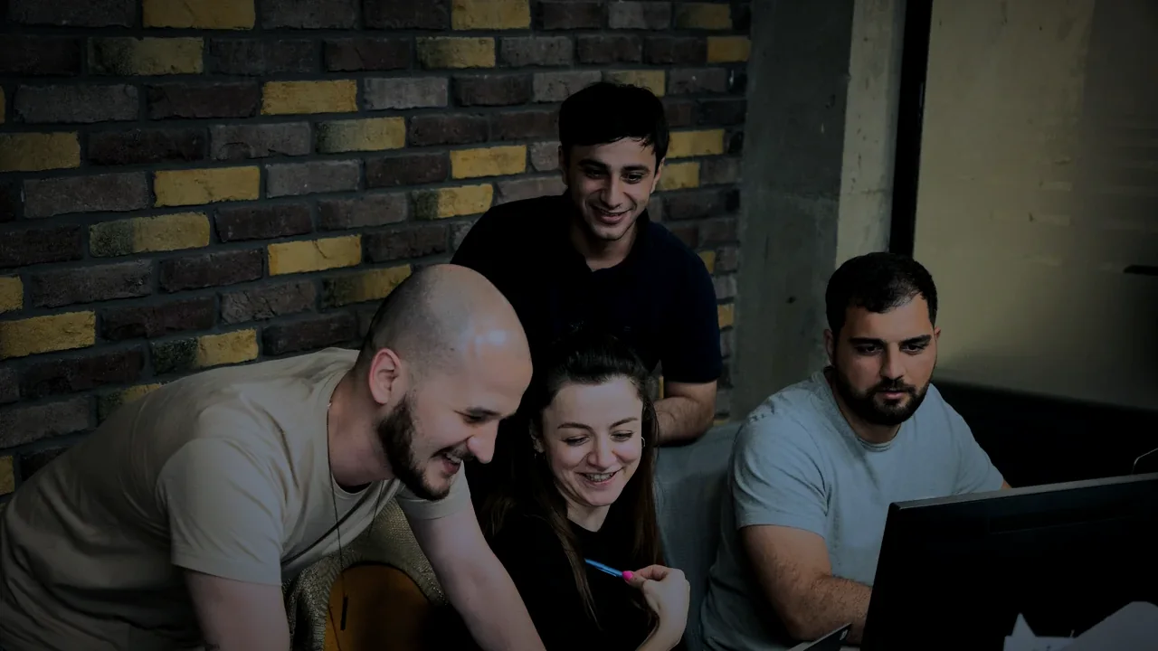 Bir ofiste tek bir ekranın başında toplanmış, gülümseyerek birlikte çalışan dört kişi